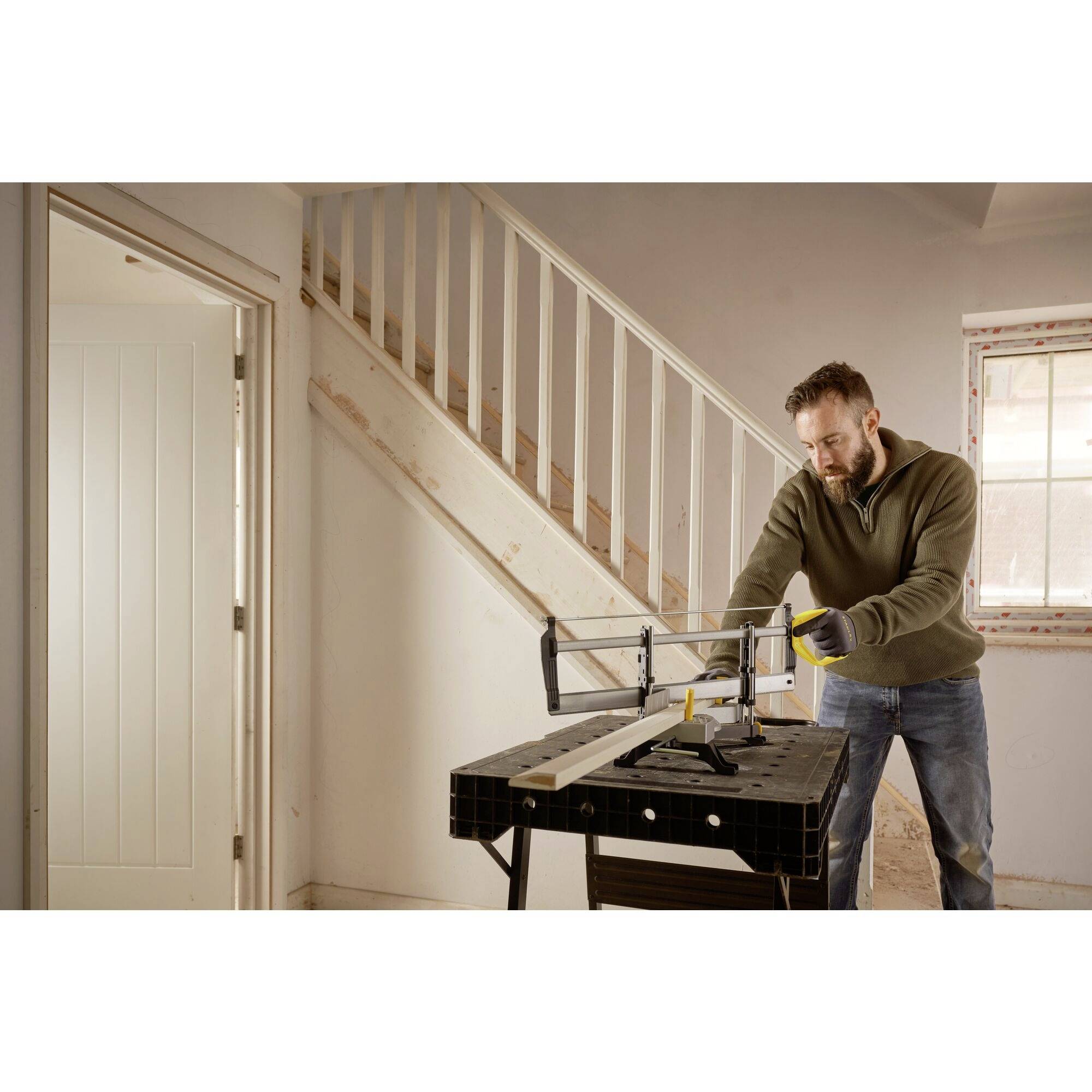A person is sawing a wooden board in a room with a staircase in the background. Light streams through a window, creating a bright atmosphere.