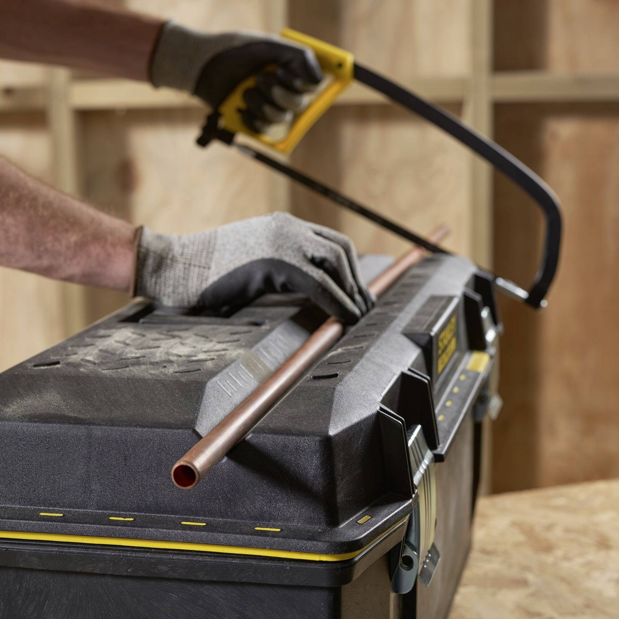A craftsman cuts a copper pipe with a metal saw on a black workbench, wearing grey work gloves.