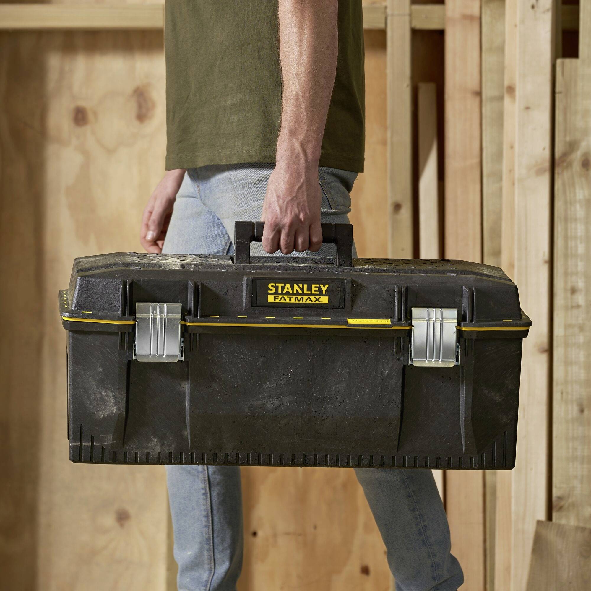 A person is carrying a black Stanley FatMax toolbox with a yellow logo while walking in a woodworking workshop.