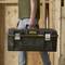A person is carrying a black Stanley FatMax toolbox with a yellow logo while walking in a woodworking workshop.