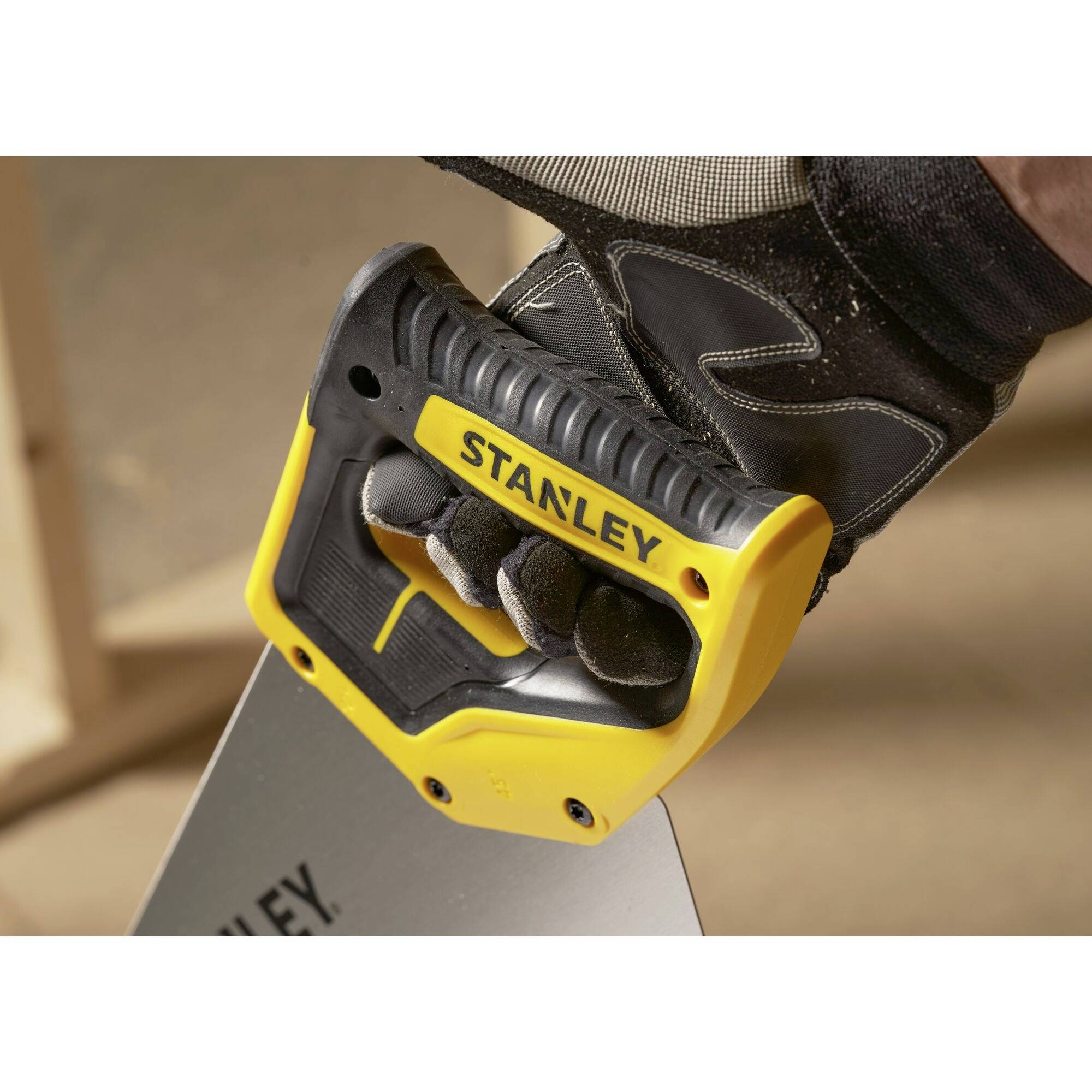 A hand is holding a fox-tail saw with a yellow Stanley handle. The background shows wood being worked on in soft focus.