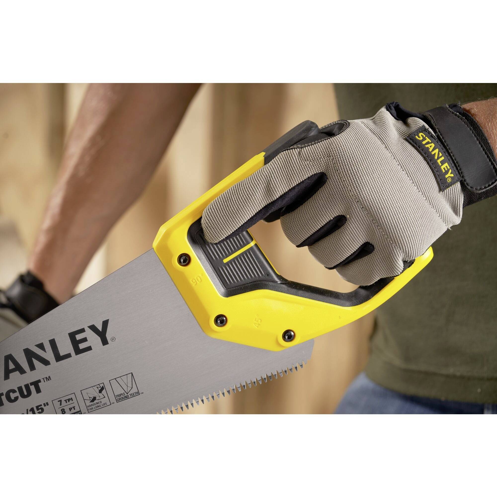 A person is holding a Stanley handsaw with a yellow handle and is wearing gloves. Blurred wooden walls are visible in the background.