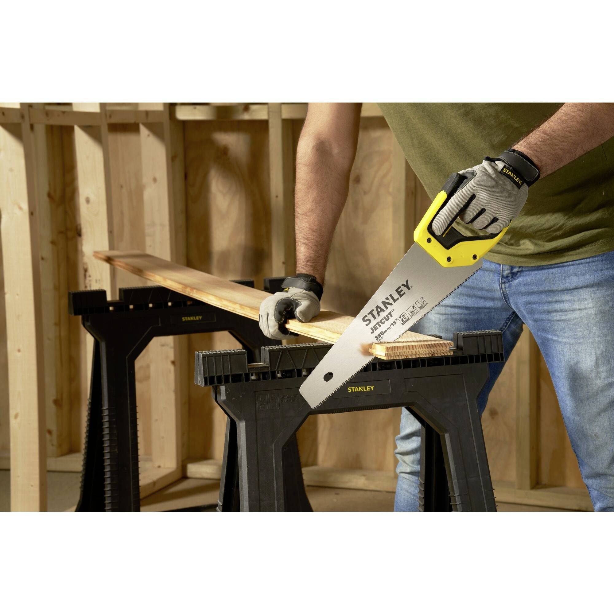 A person is cutting a wooden board with a Stanley hand saw, which is resting on two sawhorses. Hands are protected with work gloves.