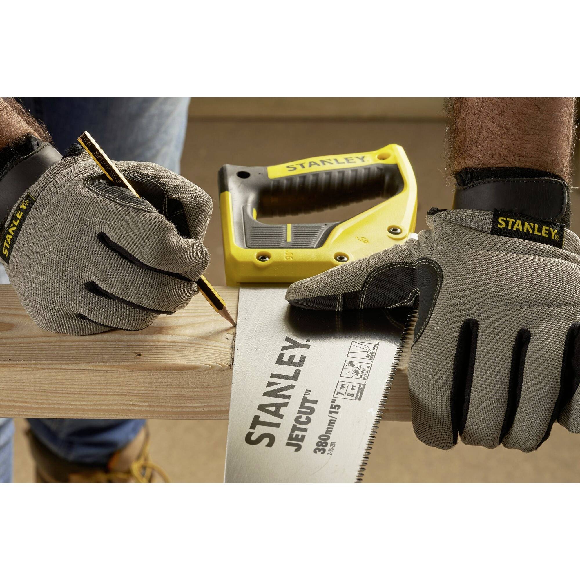 Two hands wearing work gloves draw with a pencil on wood. A yellow handsaw and measuring tape are visible beside them.