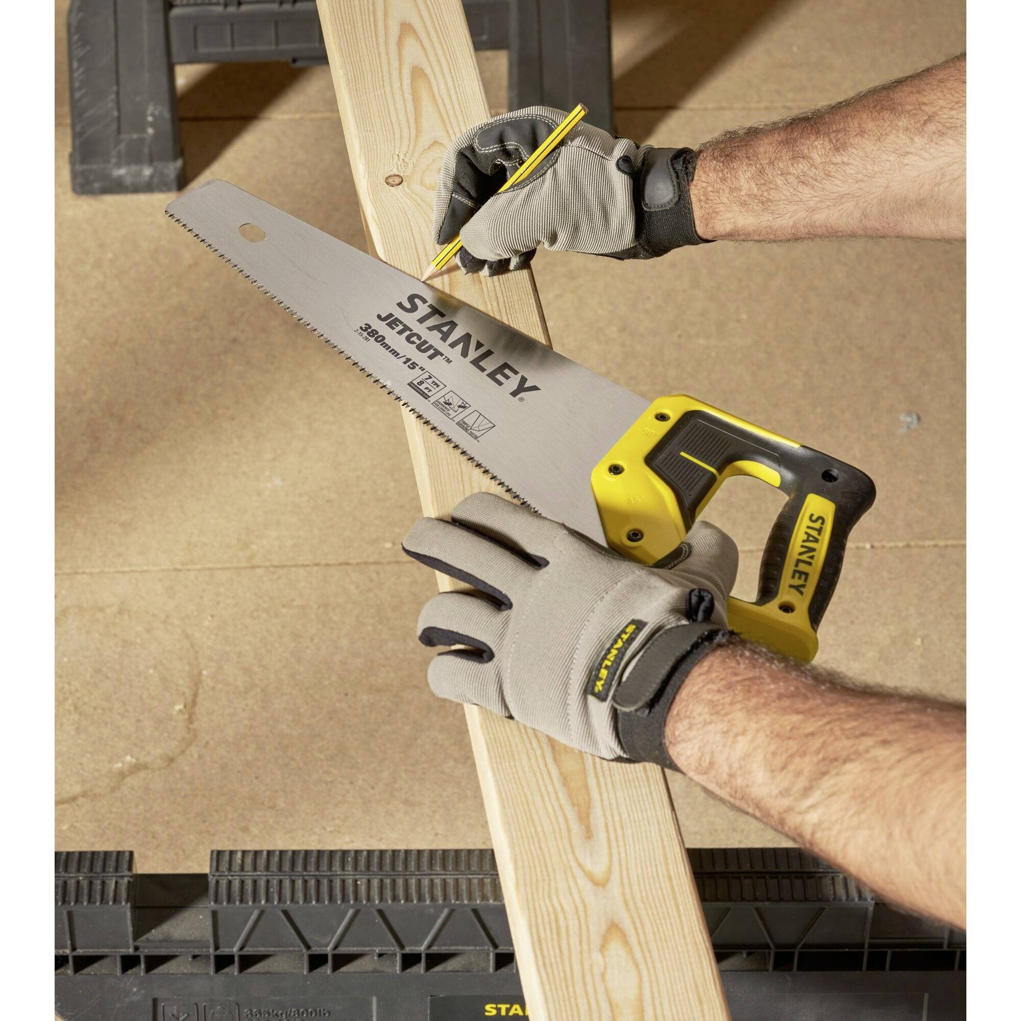 A person is sawing a wooden board with a Stanley handsaw, wearing grey gloves.