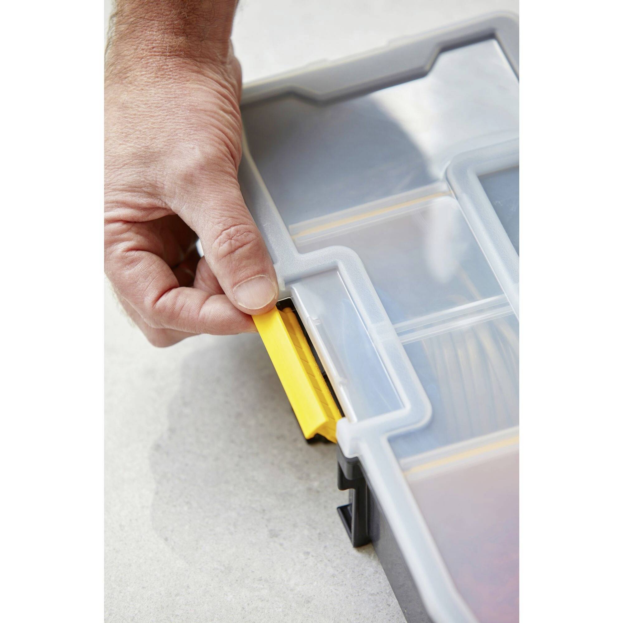 A hand opens a transparent plastic container with a yellow lid on a light background.