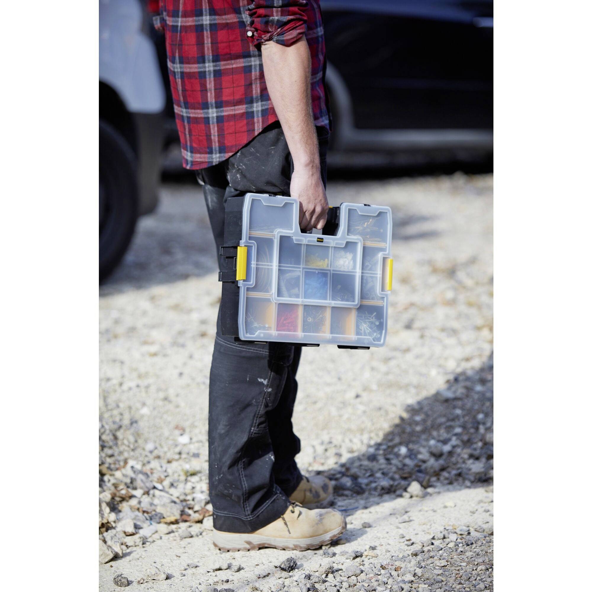 A person is carrying a transparent toolbox with a yellow lid, standing on a gravel path, partially in the shade.