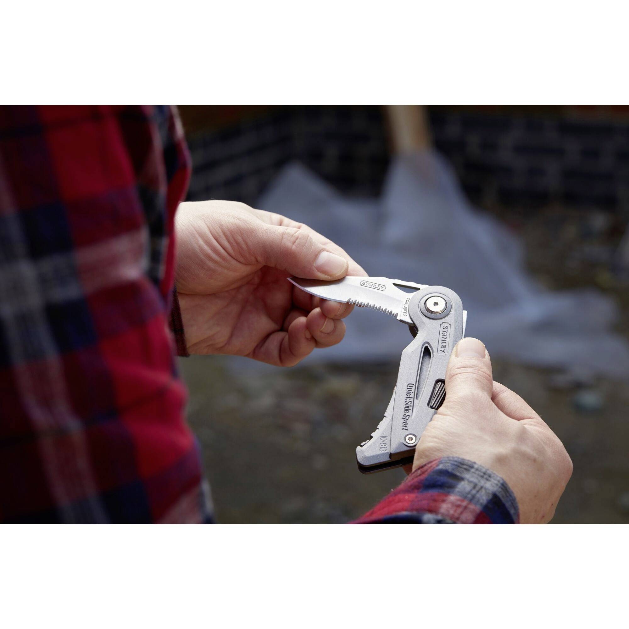 A person in a lumberjack shirt is holding a folding multi-tool while examining it. The background is blurry and shows various materials.