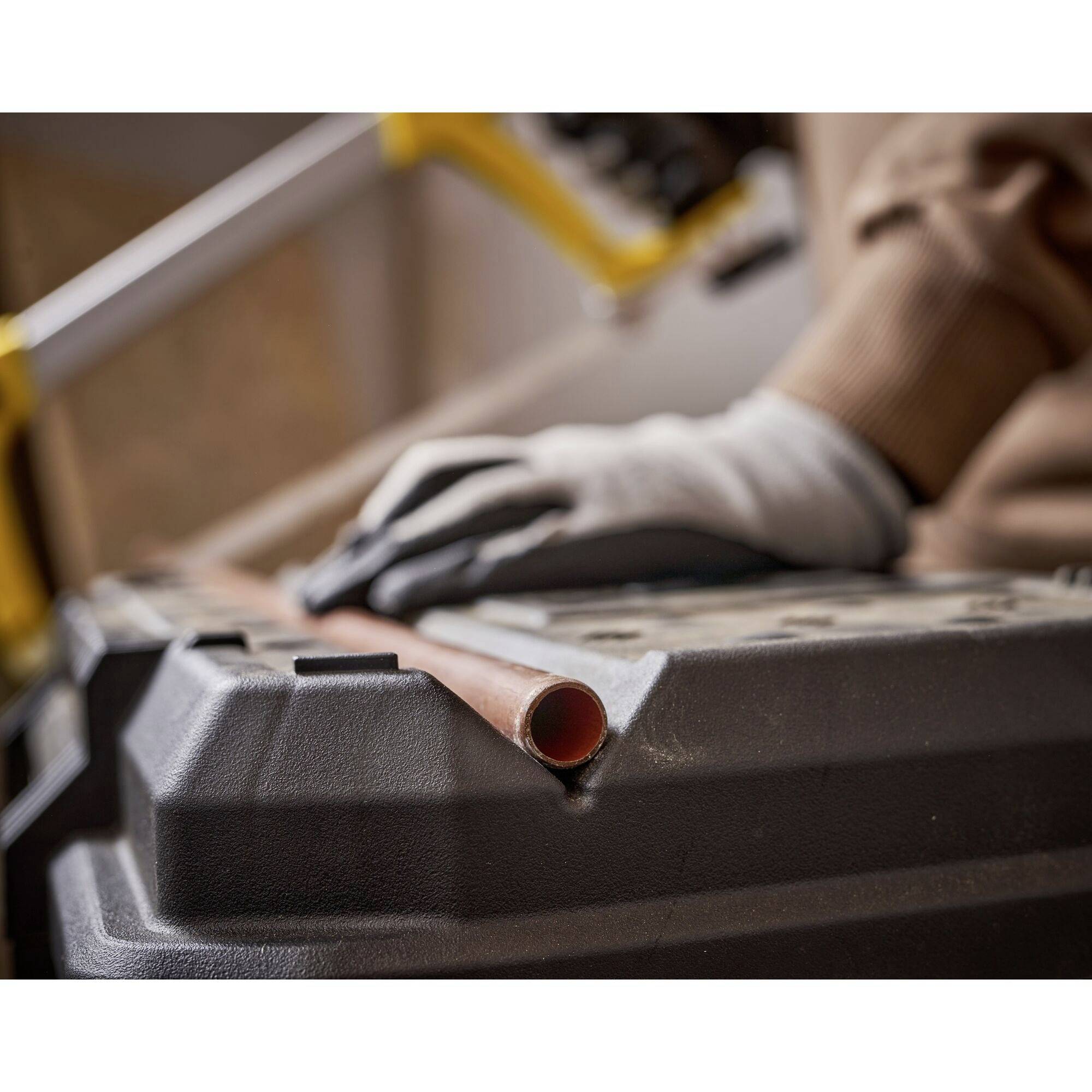 A person is cutting a copper pipe with a metal saw on a sawing trestle. They are wearing gloves for protection.