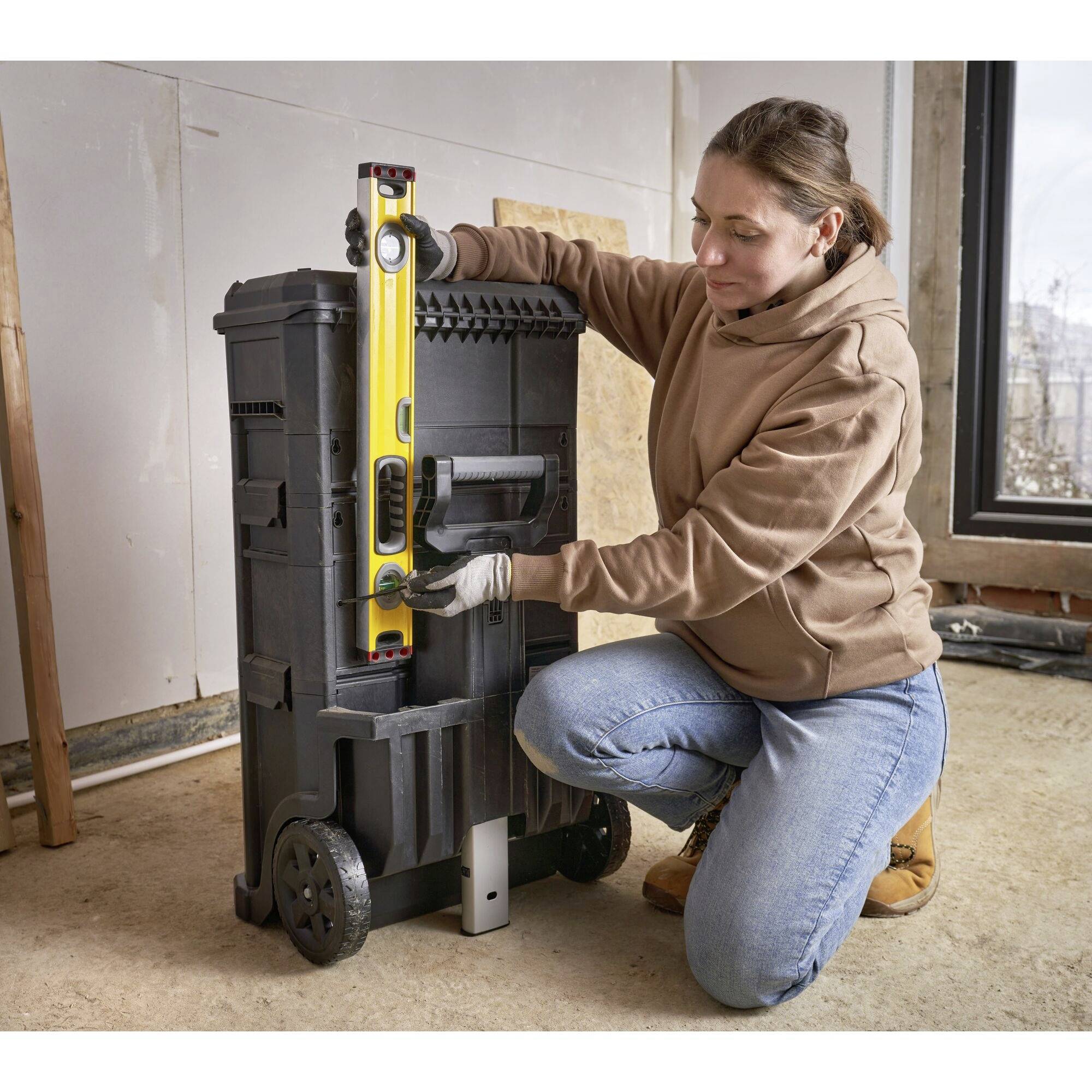 A person wearing a brown hoodie and blue jeans is kneeling beside a wheeled toolbox and aligning a yellow spirit level.