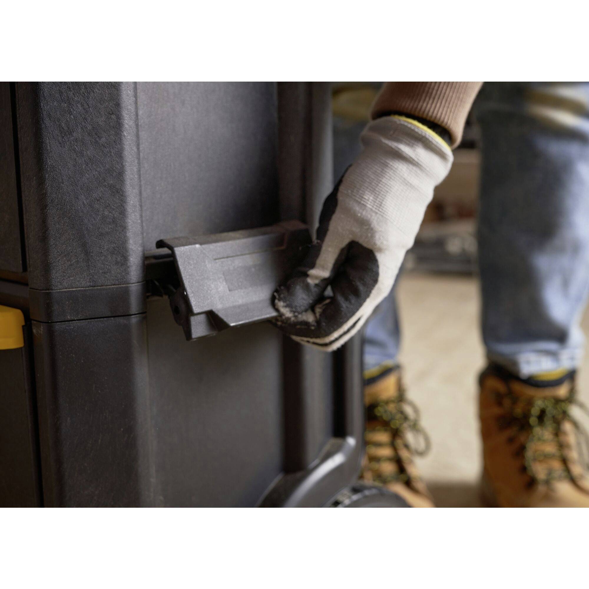 A person wearing a glove opens a compartment on a black device. They are wearing work clothing and boots.