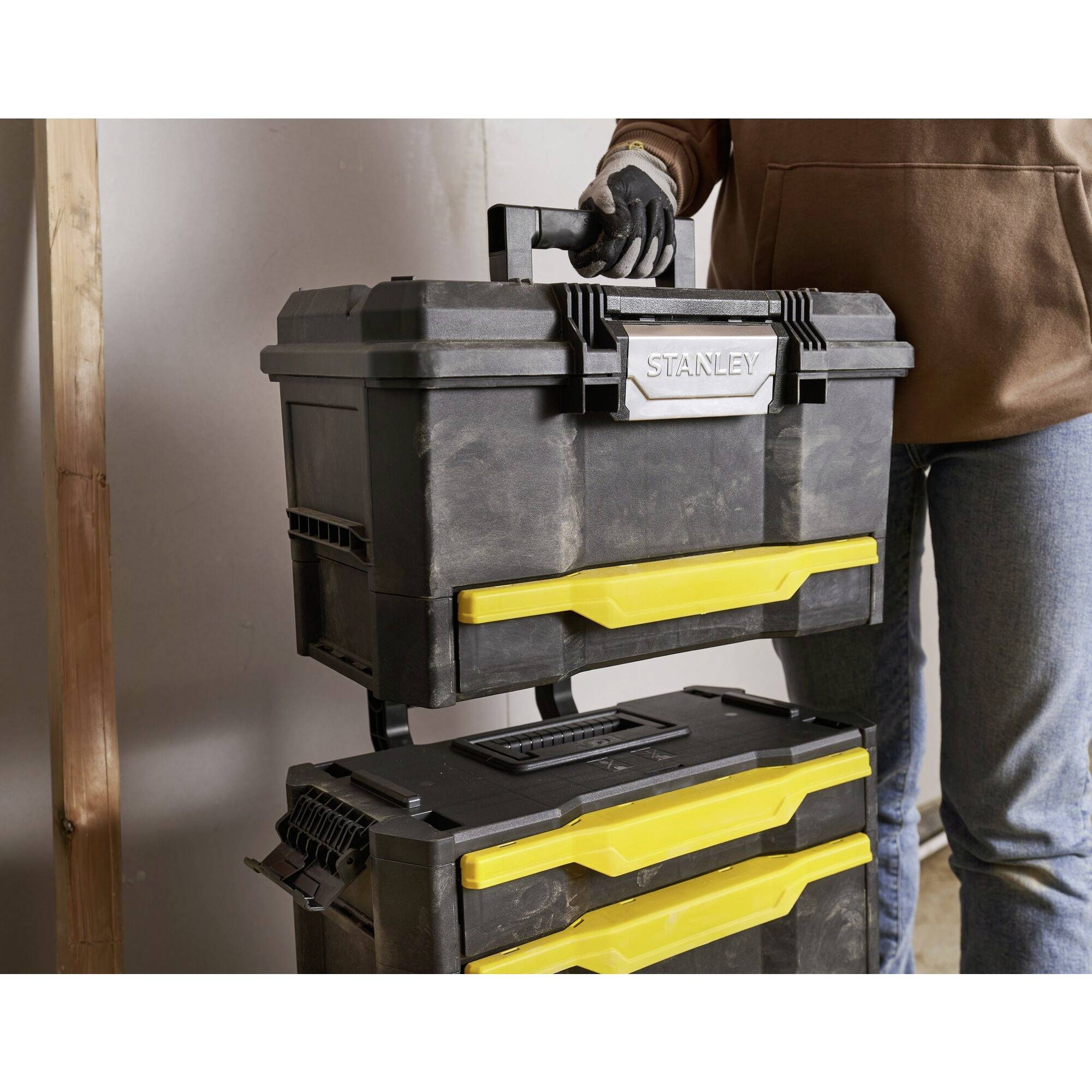 A person is lifting a black toolbox with a yellow handle, which is stacked on top of another box. The box is labelled 'Stanley'.