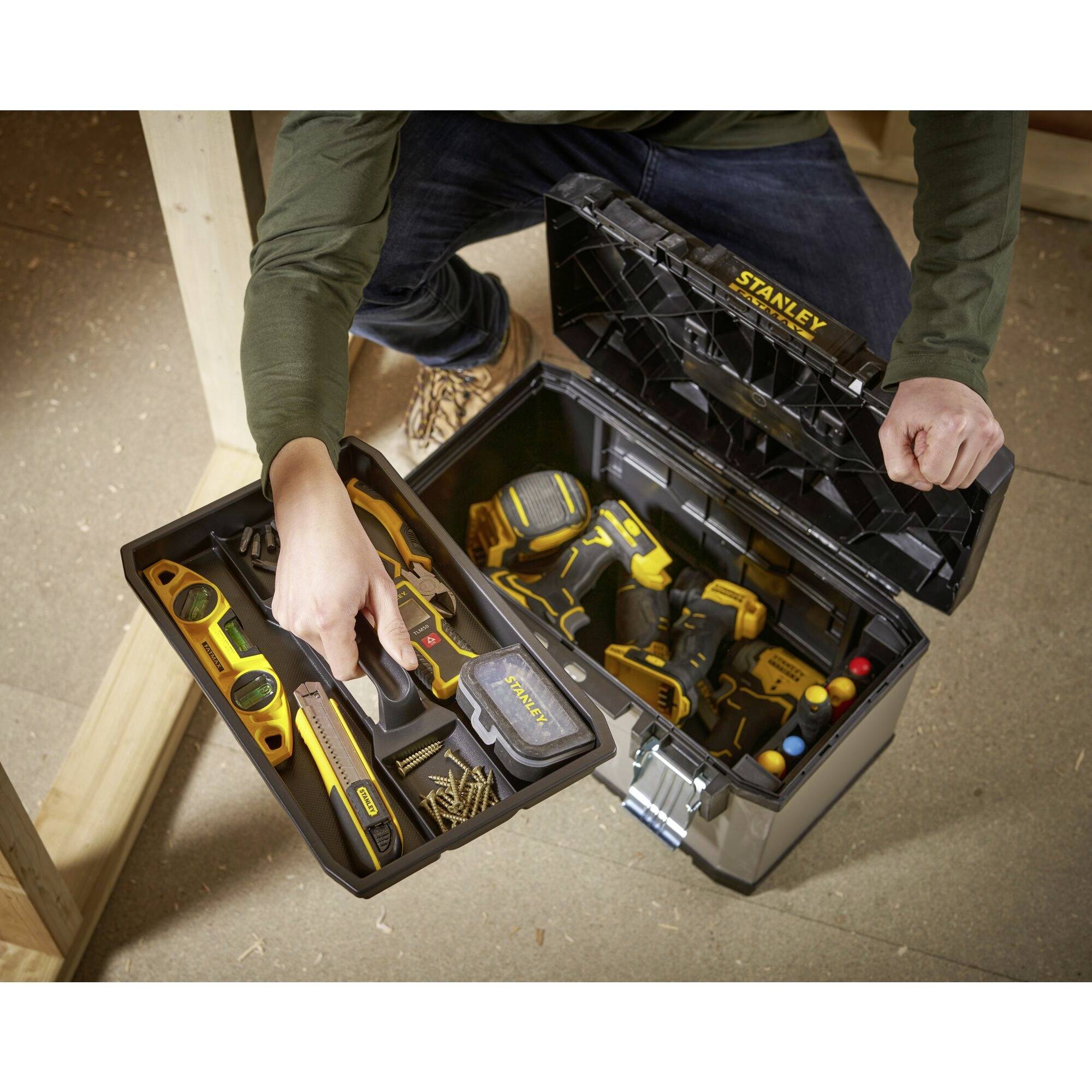 A man opens a tool case with yellow tools, including a spirit level and screwdriver, while kneeling in a workshop.