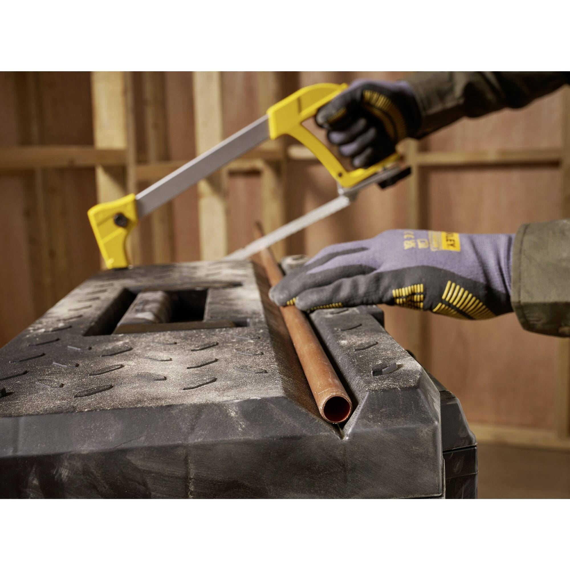 A person is sawing a copper pipe with a metal saw on a workbench. The person is wearing work gloves.