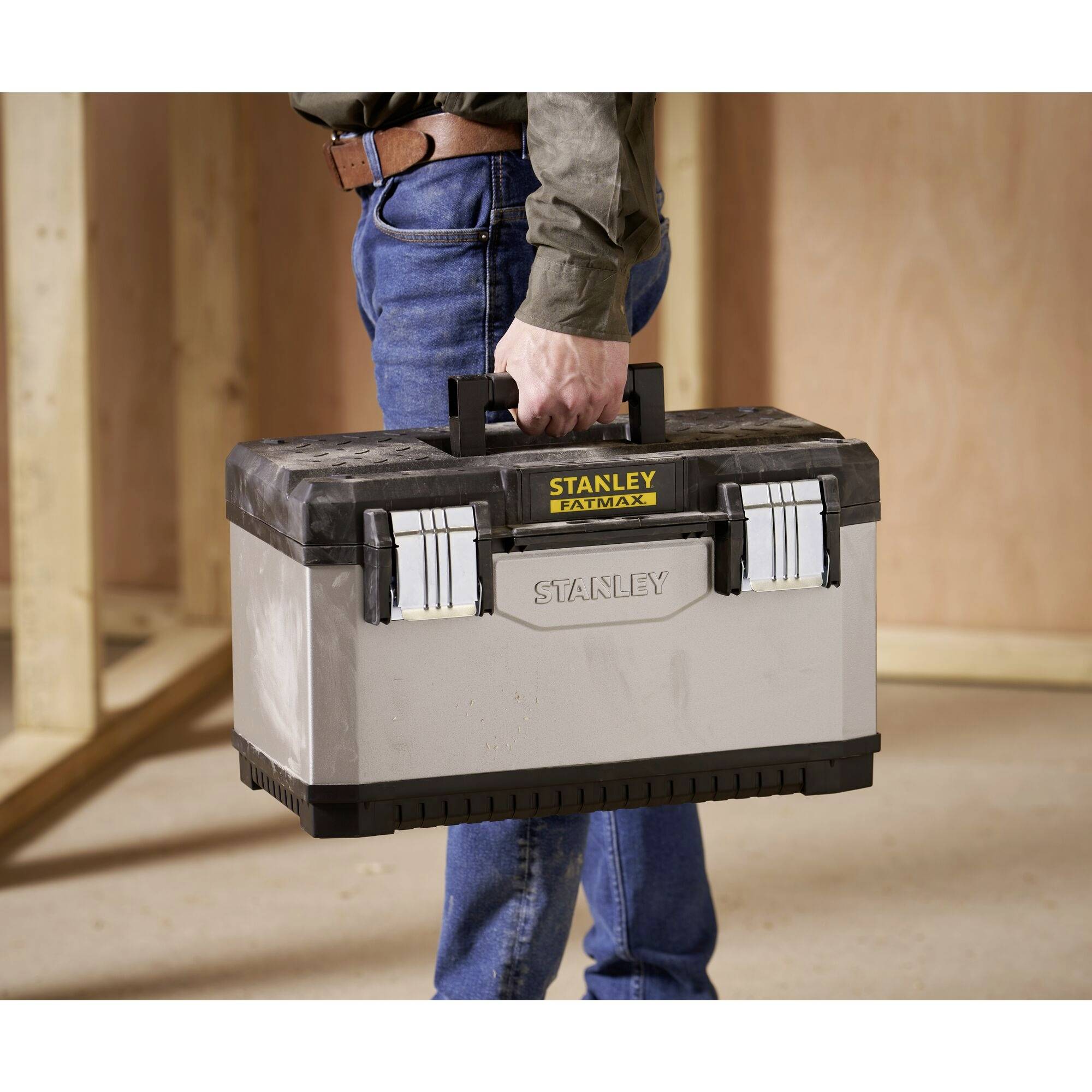 A person is carrying a grey toolbox with a black lid that reads 'Stanley Fatmax' in a construction room.