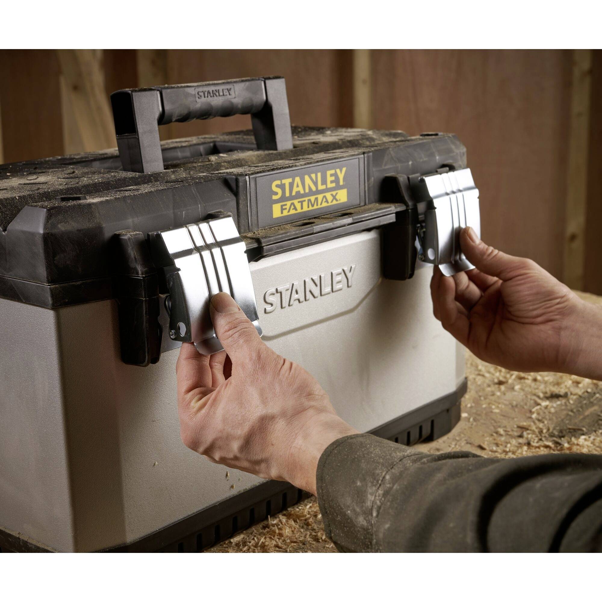 A person opens a Stanley Fatmax toolbox in a workshop environment. Hands are reaching for the latches of the case.