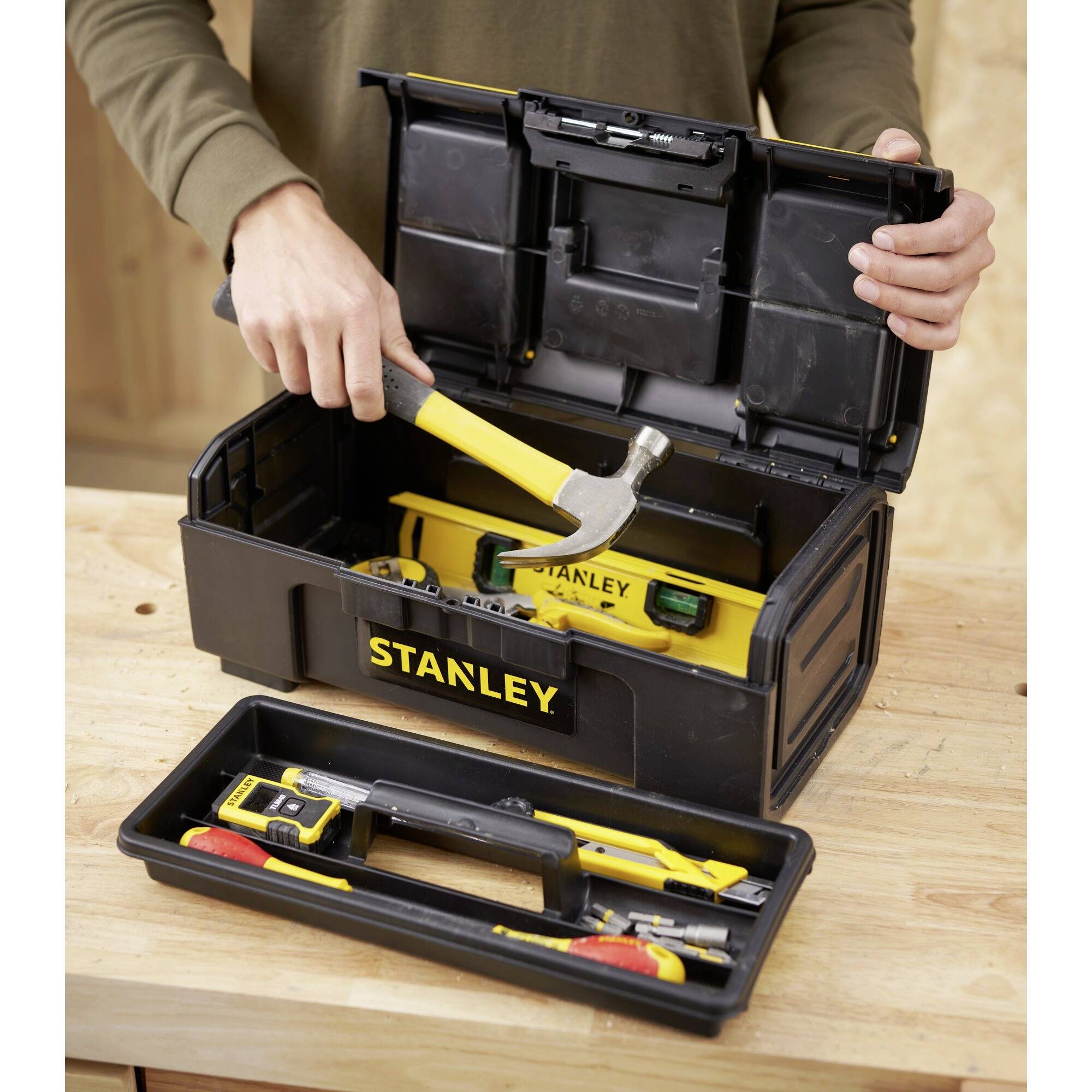 A person opens a black Stanley toolbox with a yellow hammer and spirit level on a workbench.