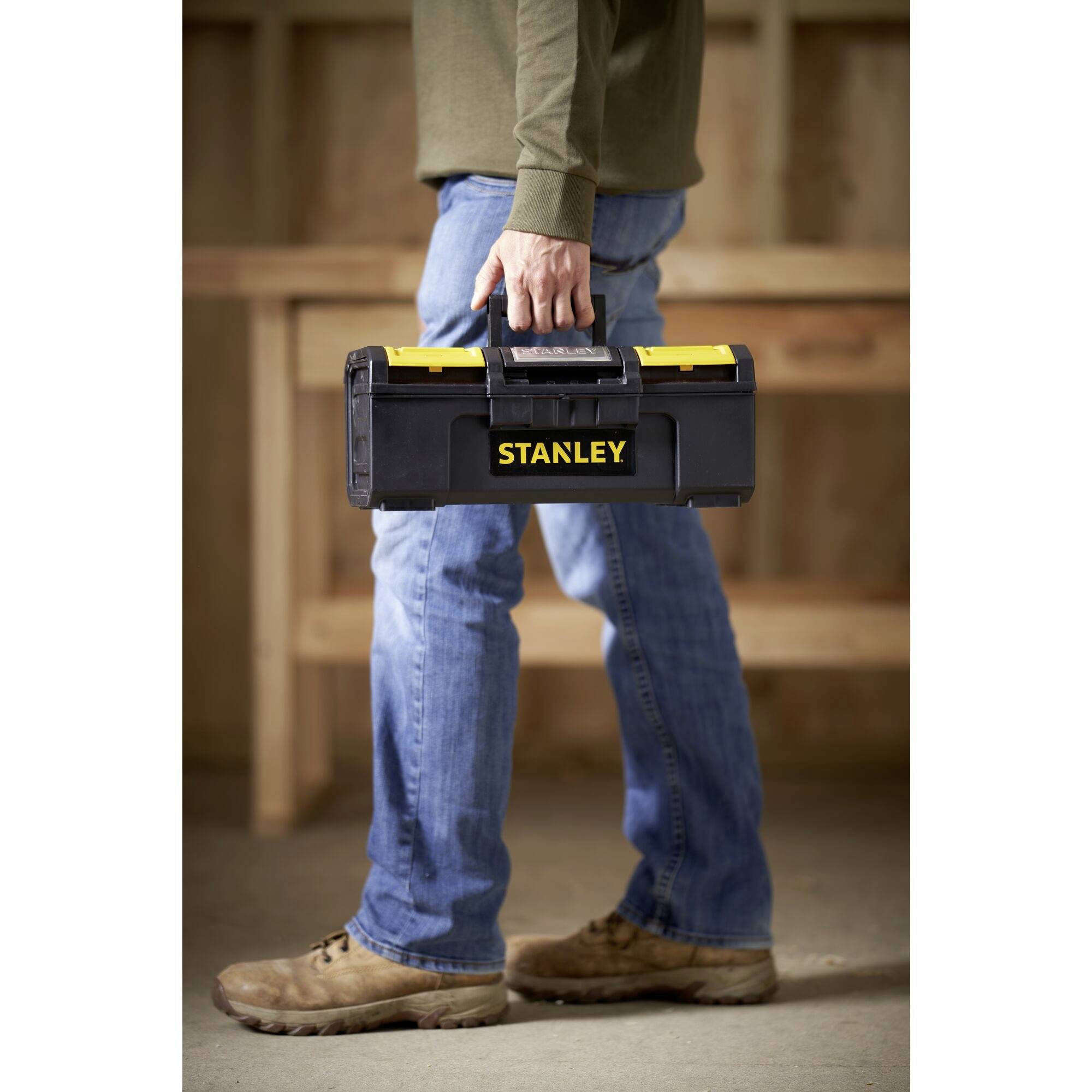 A person is carrying a black toolbox with a yellow 'Stanley' print in the workshop area.