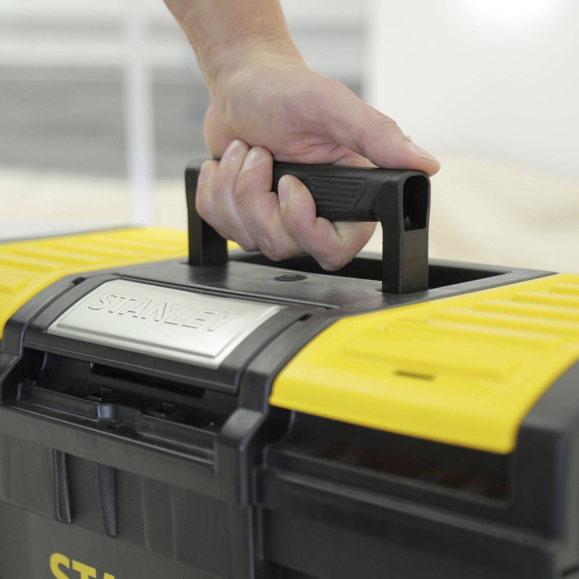 A hand lifts the handle of a yellow and black toolbox, which is standing on a surface.