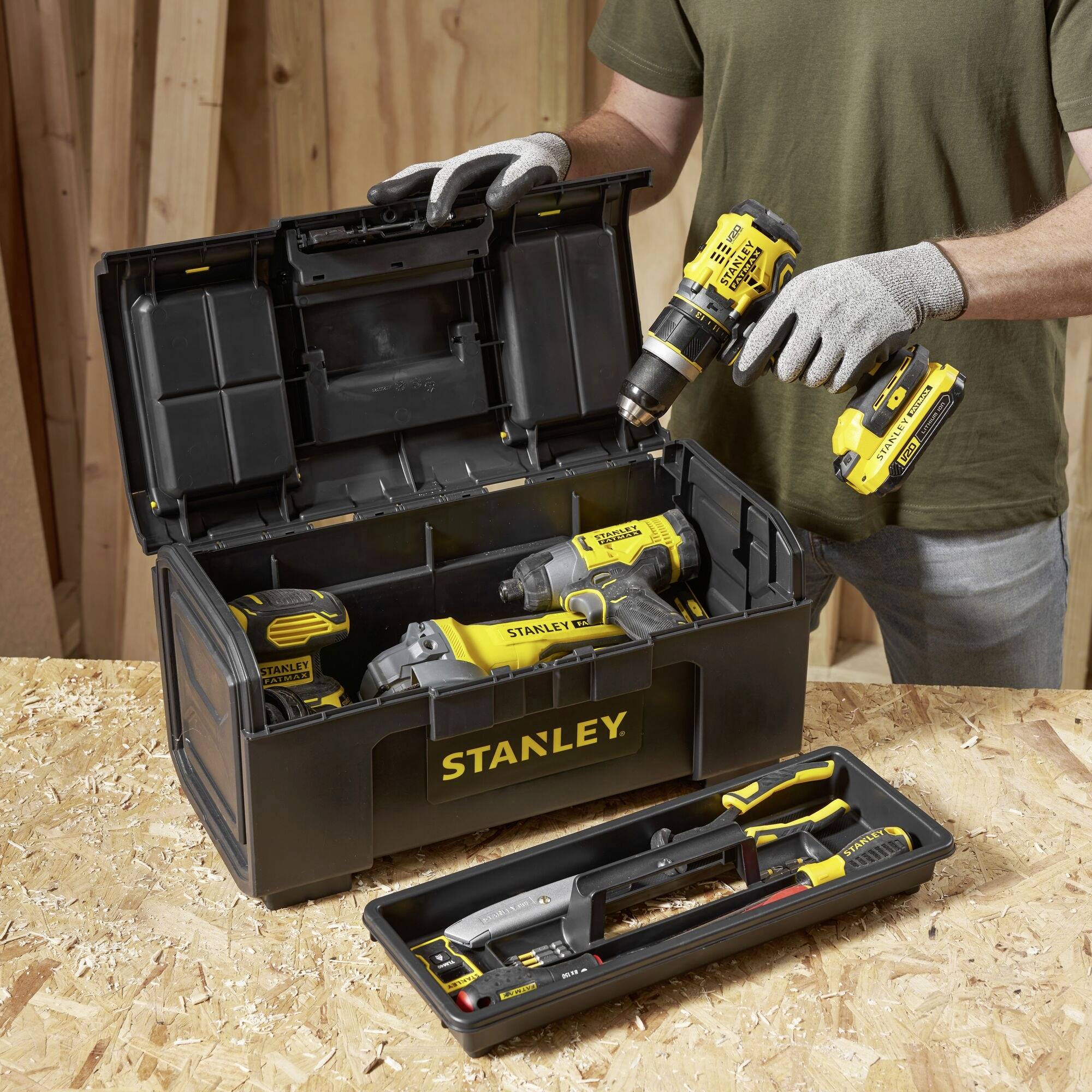 A person is holding a drill near an open toolbox with several Stanley power tools on a wooden table.