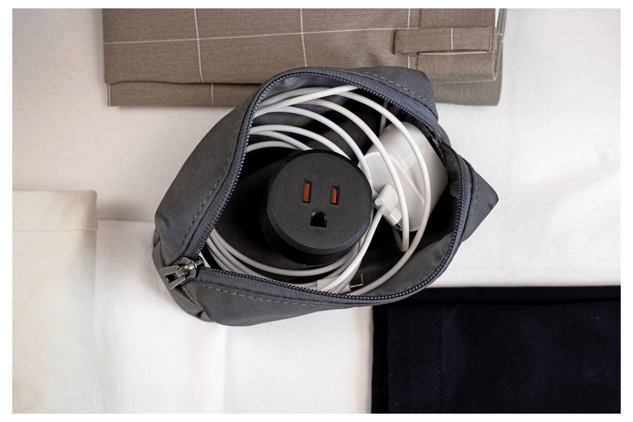Open black zip pouch holding a coiled white power cable and black electrical outlet adapter on a bed or table.