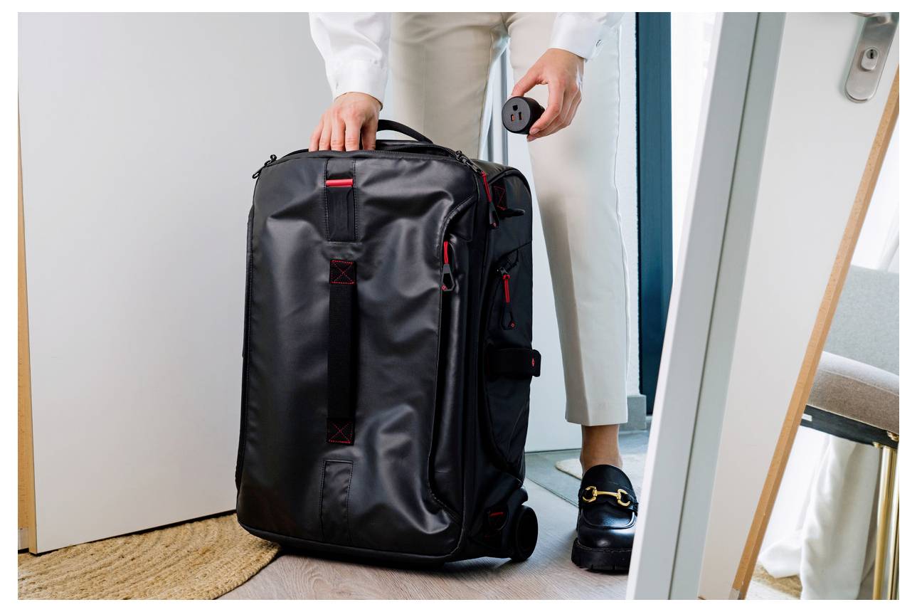 Person packing a large black wheeled suitcase in a doorway, holding the handle and a small round item, suggesting travel or departure.