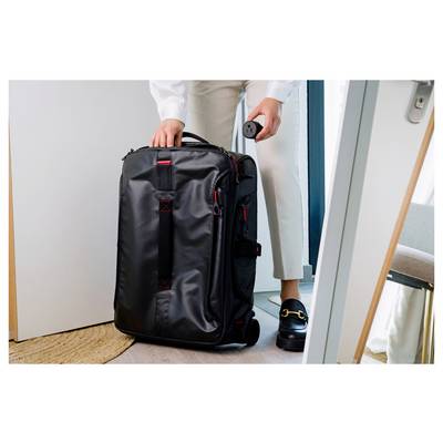 Person packing a large black wheeled suitcase in a doorway, holding the handle and a small round item, suggesting travel or departure.