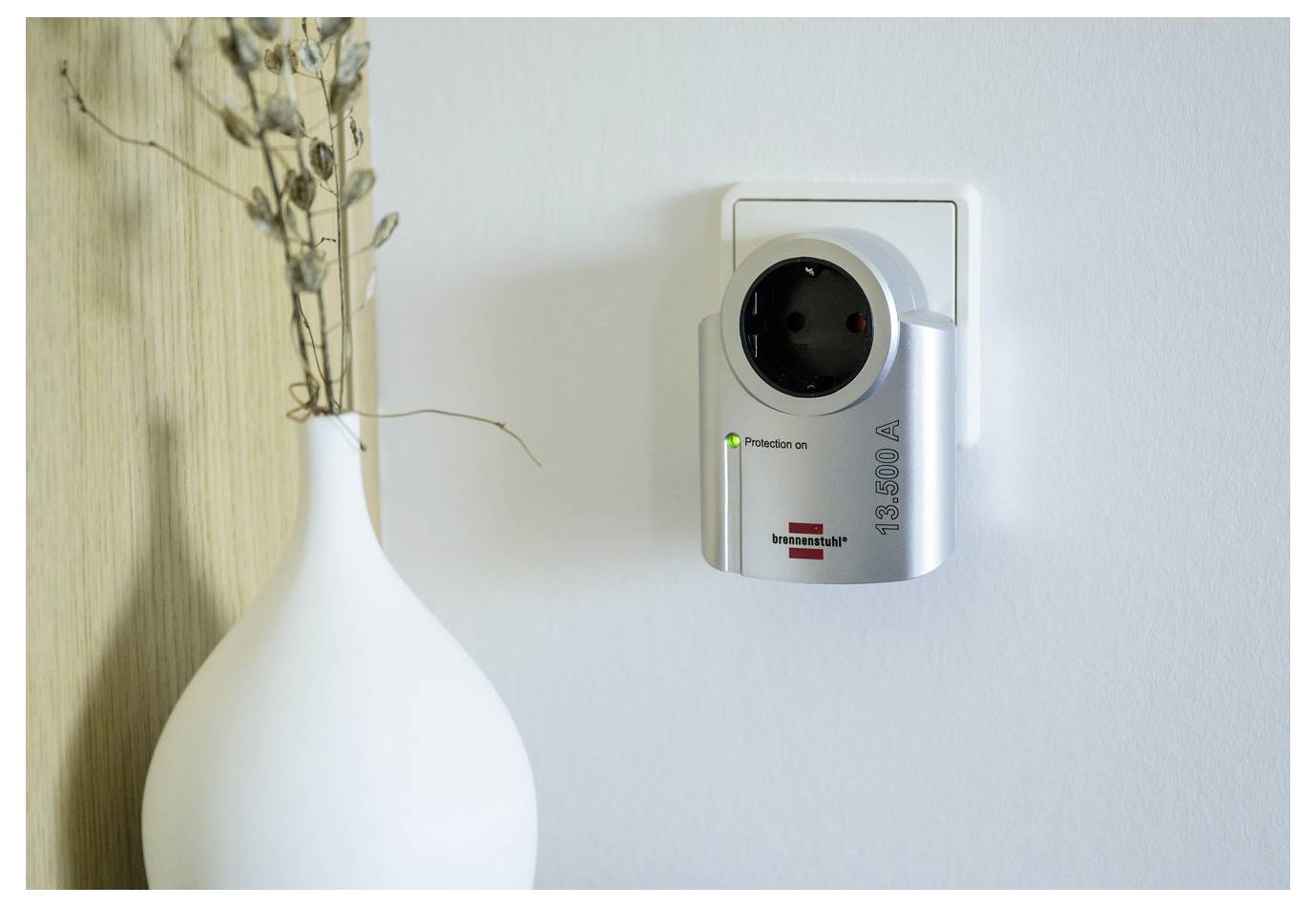 Wall-mounted air quality monitor or sensor with indicator lights beside a white vase of dried branches.<br>