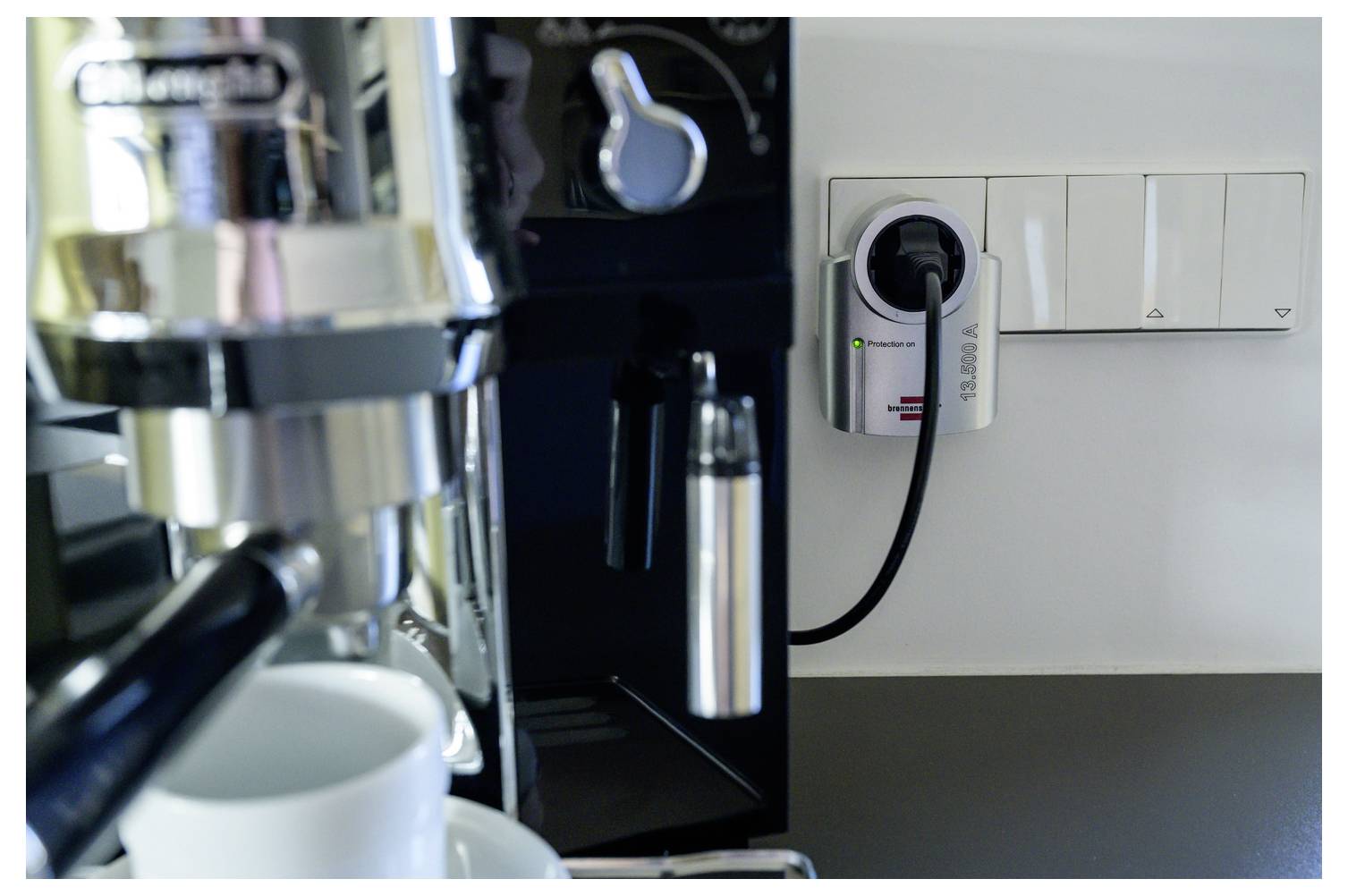Close-up of a coffee machine plugged into a wall outlet beside a light switch panel, with a cup in front.