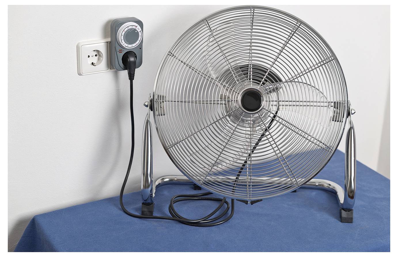 Large electric floor fan plugged into a wall outlet on a blue surface, with a control unit mounted beside the plug.