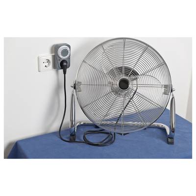 Large electric floor fan plugged into a wall outlet on a blue surface, with a control unit mounted beside the plug.