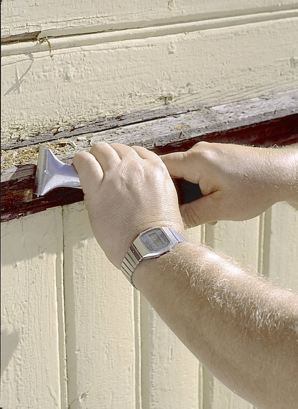 A person is removing old paint from a wooden window frame using a scraper. They are wearing a wristwatch.