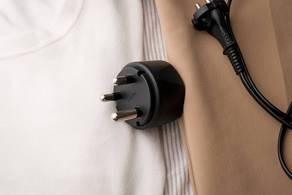 A black travel adapter with a three-pin plug is lying on beige fabric next to a black power cable.