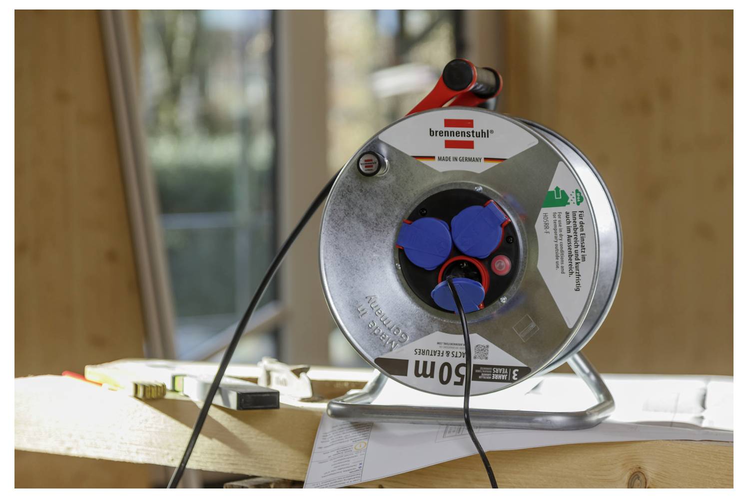A metal cable reel sits on a wooden surface in a sunlit room, with two cables plugged into its blue sockets.
