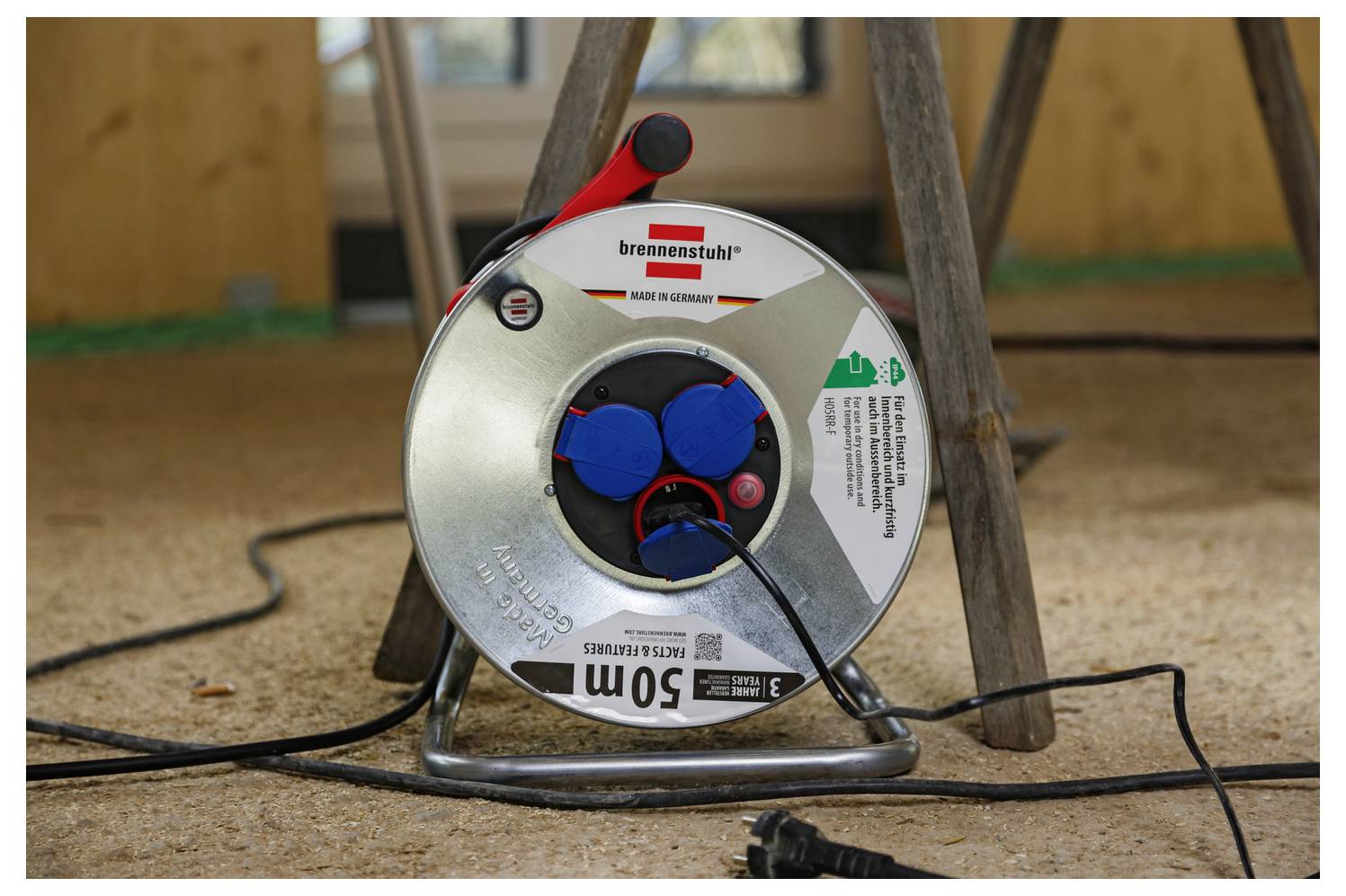 A metal cable reel with three blue sockets and a plug lies on the floor beneath a wooden structure, used for extending electrical connections.