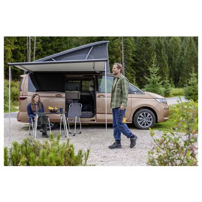 Two people camping beside a tan camper van with pop-up awning; one sits at a small table while the other walks nearby in a forest clearing.