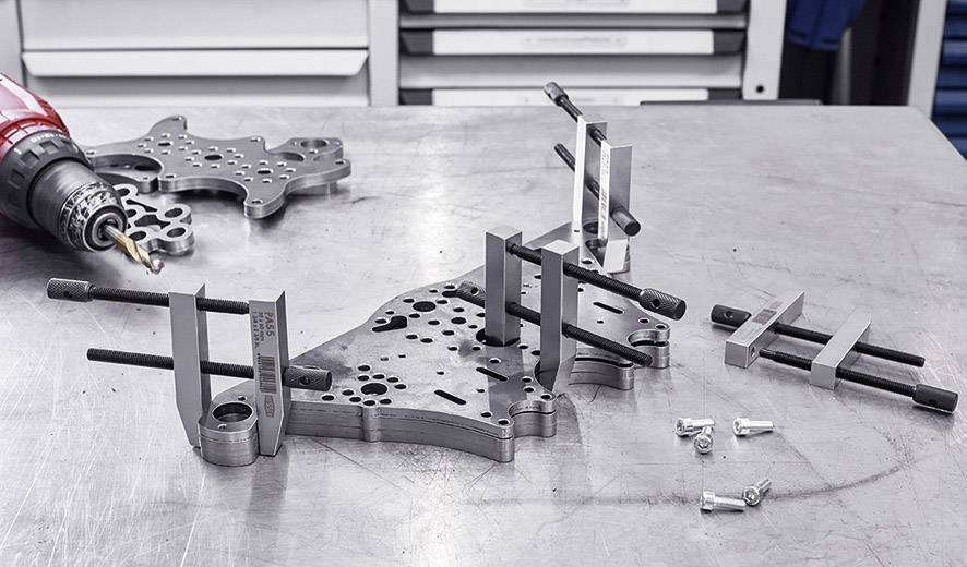 Metal parts and tools are lying on a workbench in a workshop. A drill is positioned on the left, and screws are scattered about.