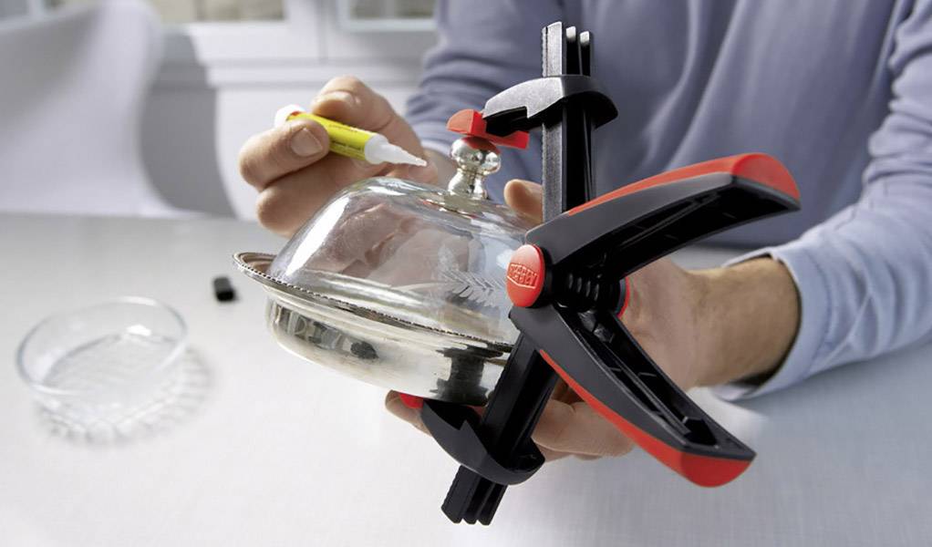 A person is repairing a broken metal lid knob with adhesive while securing the lid with a clamp.