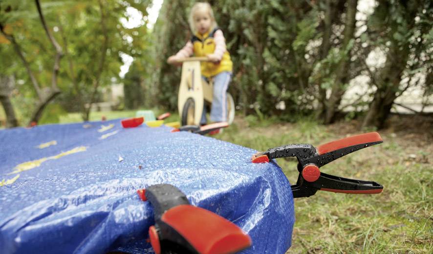 A child plays on a wooden toy in the background, while in the foreground a blue fabric is secured to a table with clips.