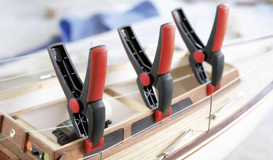 Three clamps with red handles hold together the hull of a wooden ship model during construction.