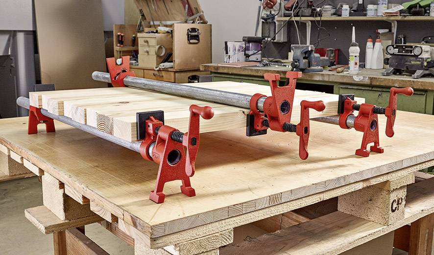 Woodworking workshop with a piece of wood clamped in red pipe clamps on a workbench. Tools and shelves visible in the background.