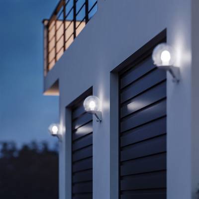 Wall lights on a modern building illuminate a terrace at dusk. The sky is overcast, and vegetation can be seen in the background.