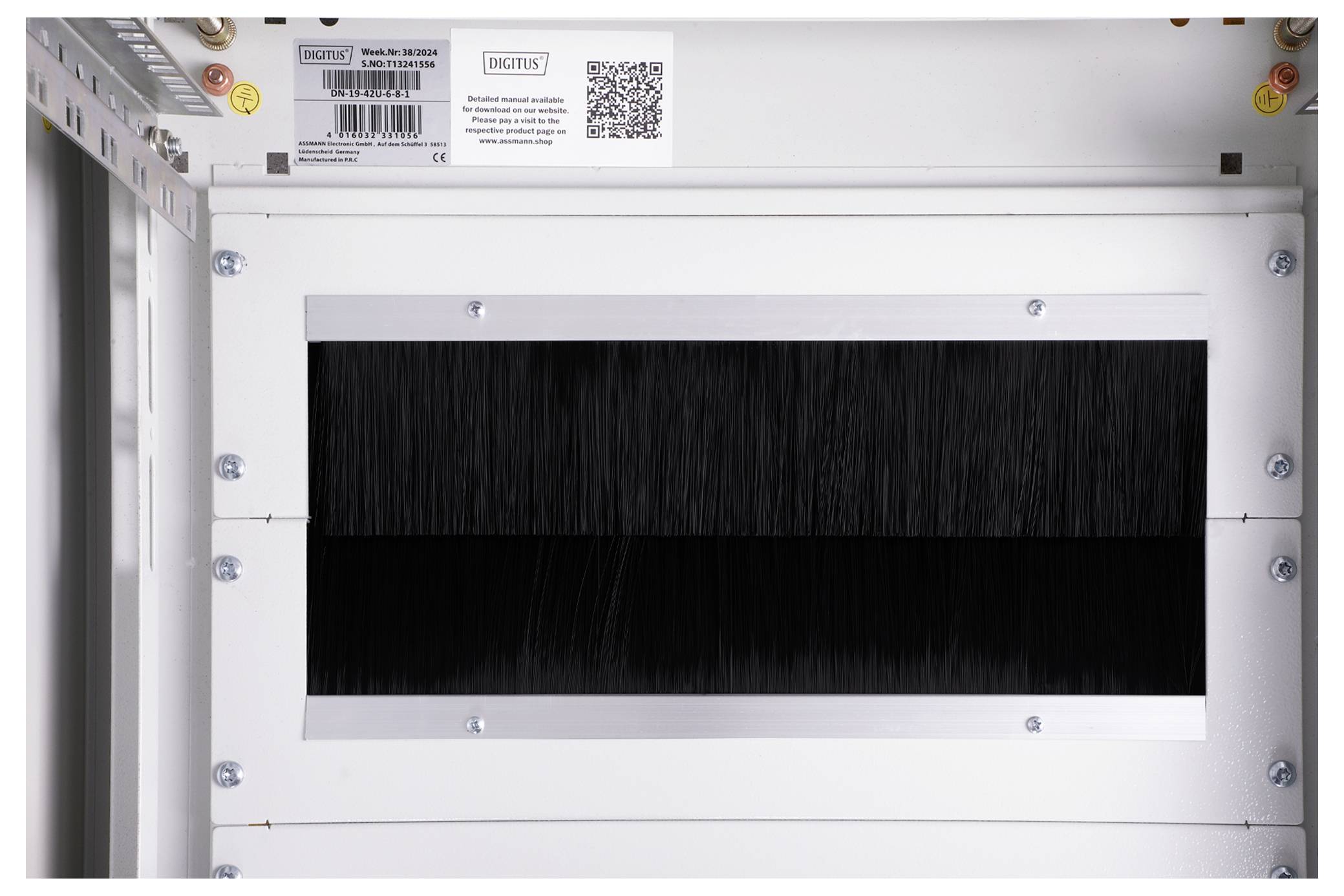 'White industrial wall with a rectangular ventilation grille. Above it, a QR code and label with technical information and barcode.'