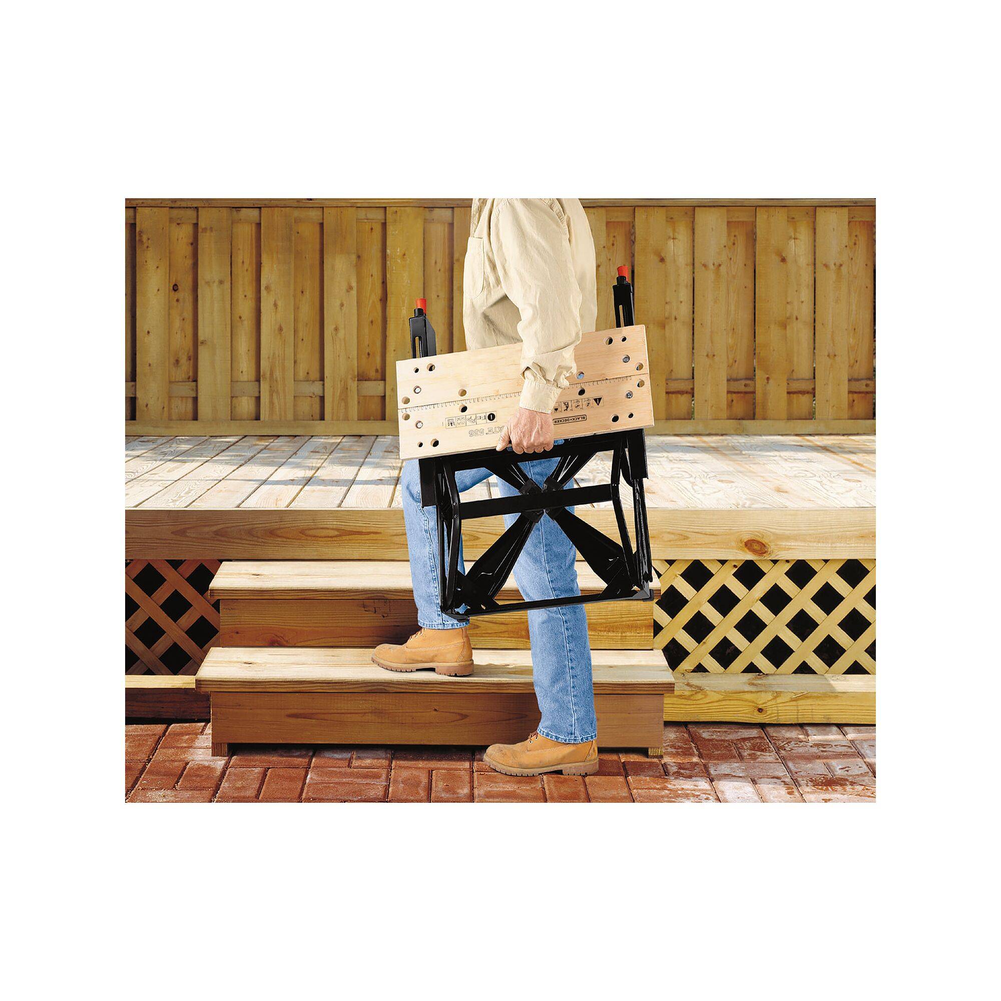 A person is carrying a folded wooden and metal folding chair up a wooden staircase. A wooden veranda is visible in the background.