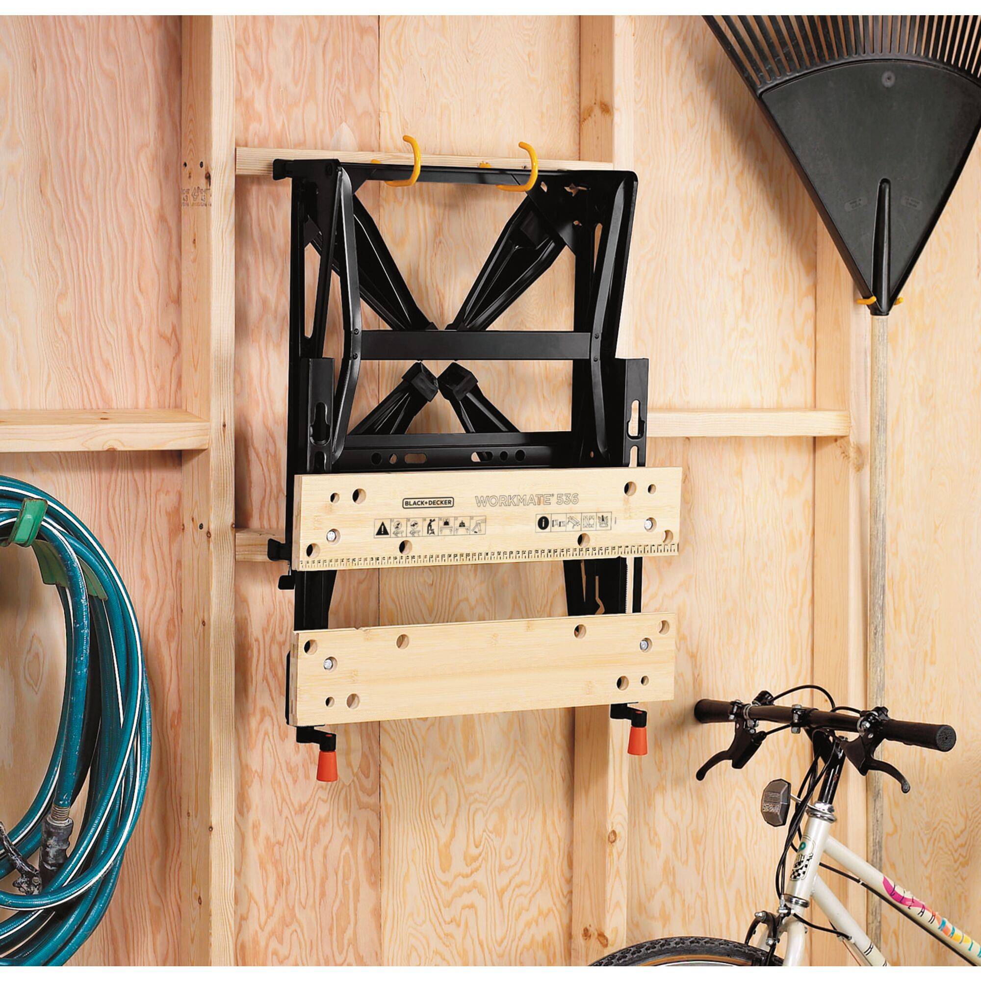 A folded workbench hangs on the wall of a wooden shed. Next to it are a garden hose, a rake, and a bicycle handlebar.