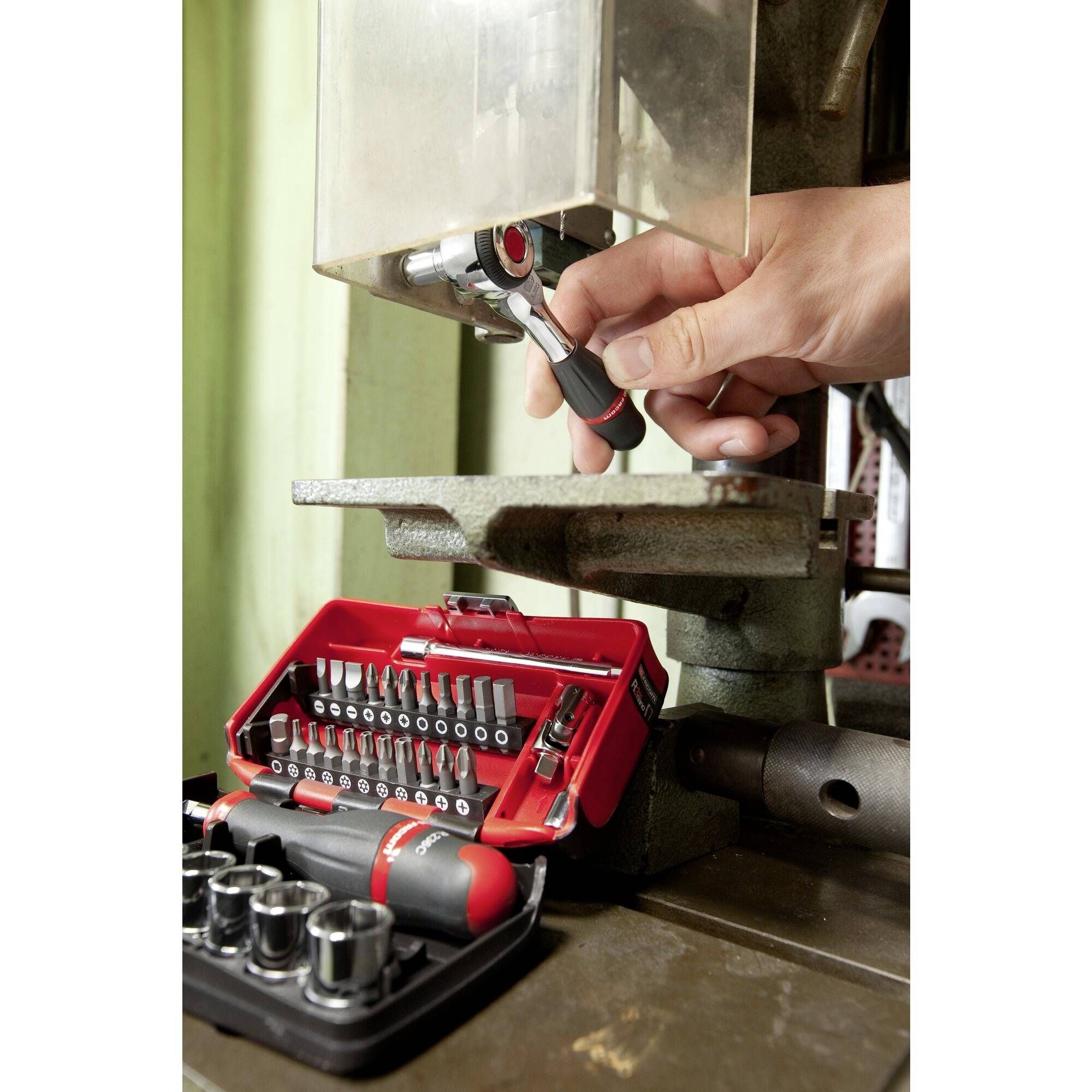 A hand adjusts a metal drill bit on a machine, close to an open red toolbox containing various metal attachments.