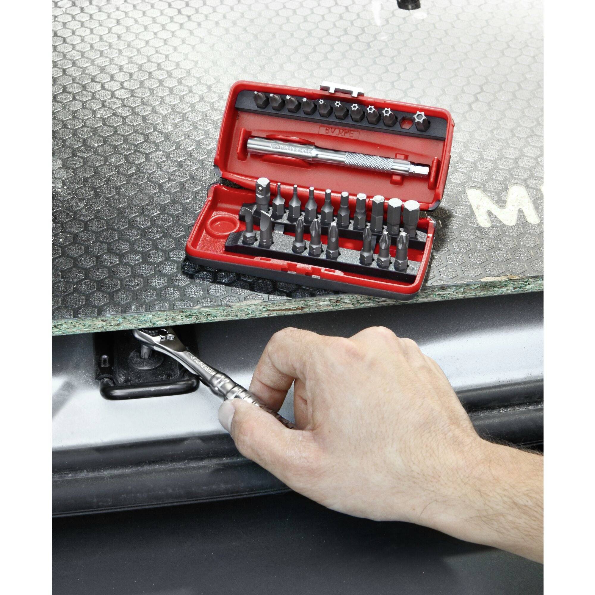 A hand holds a screwdriver and removes a screw from a car part. In the background, an open toolkit is visible.