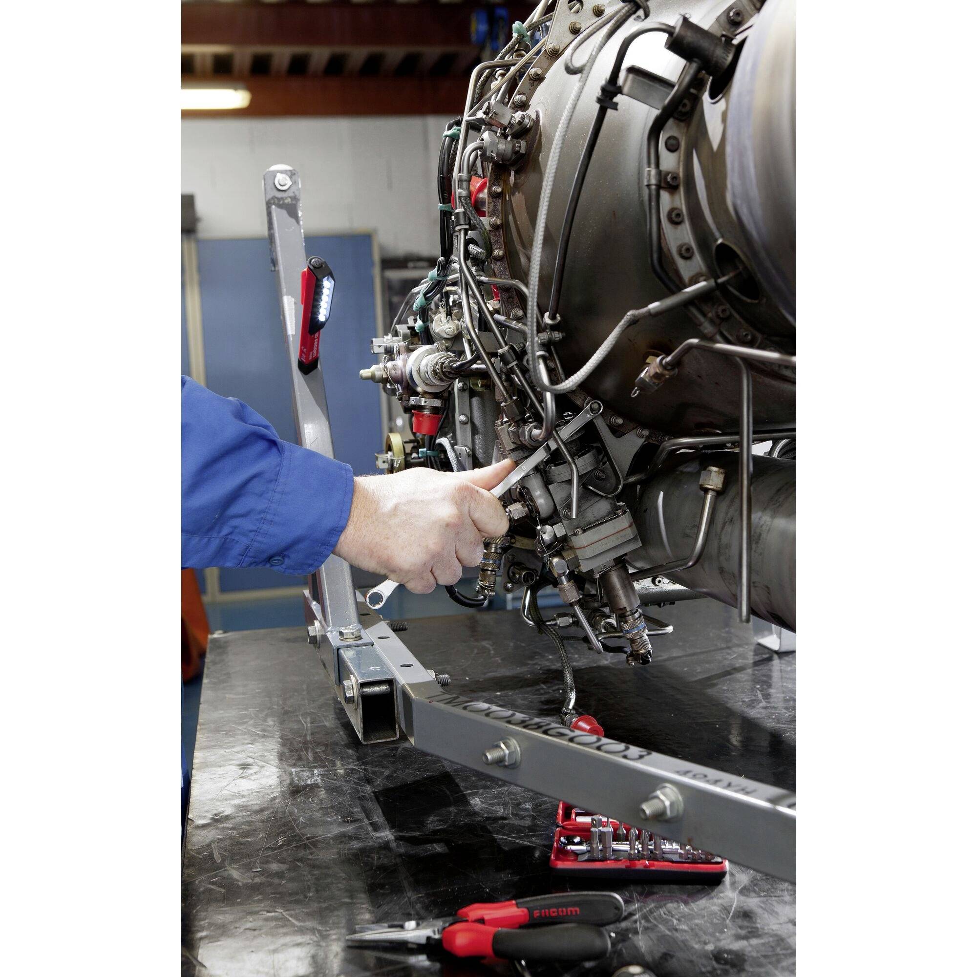 A technician in blue overalls is pointing to components of a complex engine in a workshop environment. Tools are laid out nearby.