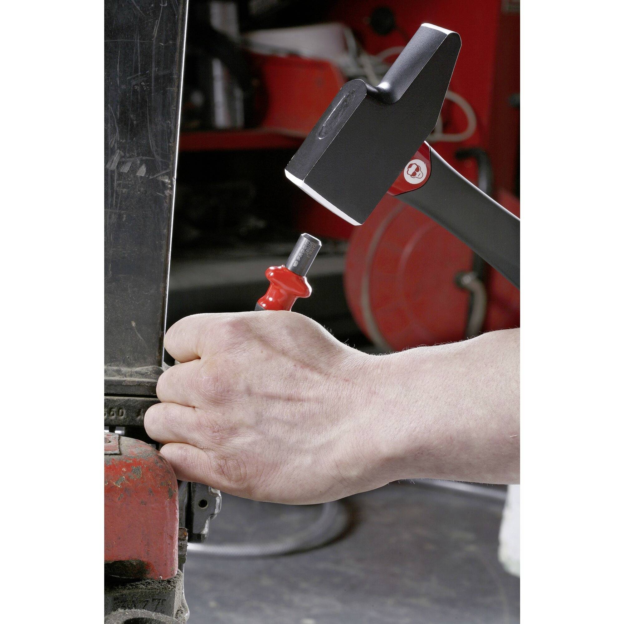 A hand is using a hammer to drive a nail into a piece of metal. Tools and a red wall are visible in the background.