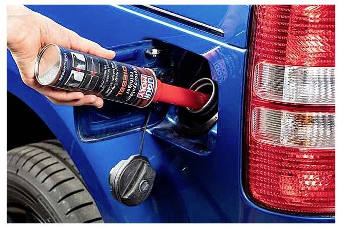 A hand pours diesel fuel conditioner from a bottle into a blue vehicle’s fuel filler opening beside the rear taillight.